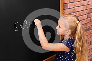 Cute little left-handed girl doing sums on chalkboard near  wall