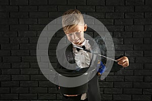 Cute little magician showing trick with hat against dark brick wall