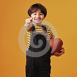 Cute little mixed race boy smiling and pointing while holding a football and posing against an orange copyspace