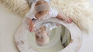 A cute little newborn baby is playing with a mirror at home lying on the bed.