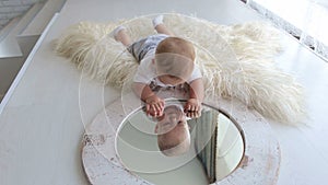 A cute little newborn baby is playing with a mirror at home lying on the bed.