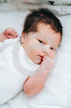Cute little newborn baby with short dark hair in white bodysuit look sleepy at camera clenching fists.