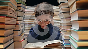 Cute little pupil boy in glasses reading interesting book in library between stacks of books literature. Education