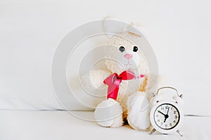 Cute little rabbit doll lying alone with blurred analog alarm clock  on white bed