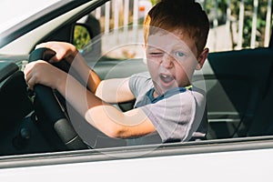 Cute little red-haired boy sitting in front of the car holding steering wheel. Boy looking at the camera and smiling