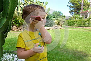 Cute little redhead boy in a green shirt listens on the phone to a voice message in the garden, outdoors