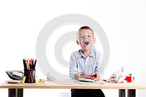 Cute little school boy with huge smile sitting at his desk on white background. Happy intelligent children in shirt with blue eye
