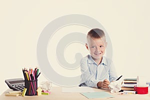 Cute little school boy with huge smile sitting at his desk on white background. Happy intelligent children in shirt