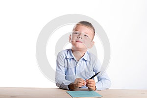 Cute little school boy with huge smile sitting at his desk on white background thinking about something