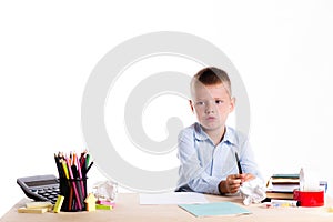 Cute little school boy with sad face sitting at his desk on whit
