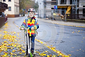 Cute little school kid boy riding on scooter on way to elementary school. Child with safety helmet, school bag on rainy