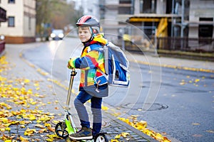 Cute little school kid boy riding on scooter on way to elementary school. Child with safety helmet, school bag on rainy