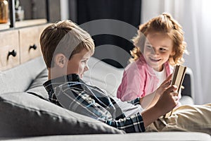 cute little schoolboy reading book on couch while his sister talking