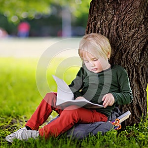 Cute little schoolboy sitting under tree and studying . Back to