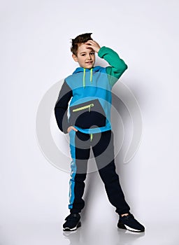Cute little schoolboy with stylish hairdo in bright cotton sportsuit stands smiling, putting hand up to his forehead.