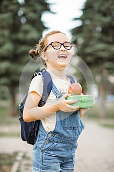 Cute little schoolgirl with backpack holding in a hands lunch box and apple. Near by school outdoor. Food for kids