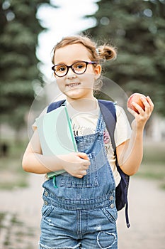 Cute little schoolgirl with backpack holding in a hands notebook and apple. Near by school outdoor