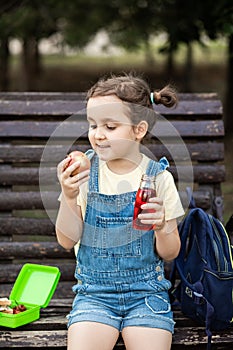 Cute little schoolgirl eating from lunch box outdoor sitting on a bench. Food for kids