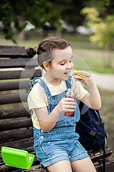 Cute little schoolgirl eating from lunch box outdoor sitting on a bench. Food for kids