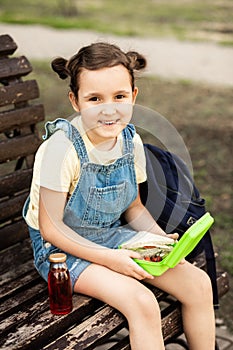 Cute little schoolgirl eating from lunch box outdoor sitting on a bench. Food for kids