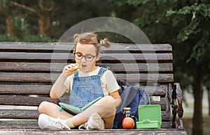 Cute little schoolgirl eating from lunch box outdoor sitting on a bench. Food for kids