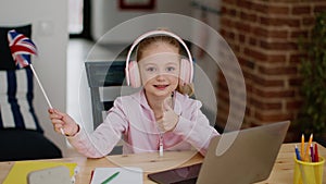 Cute little schoolgirl in headphones enjoying lessons on laptop, gesturing thumb up and waving with GB flag