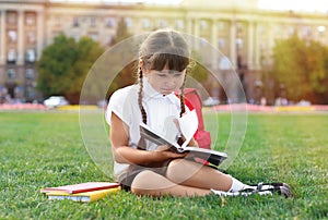 Cute little schoolgirl sitting on green grass and reading book