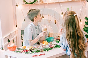 Cute little sister with long hair and an older brother drink cocoa and eat gingerbread cookies by the window