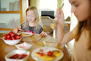 Cute little sisters enjoying their breakfast at home. Pretty children eating pancakes with strawberry sauce before school