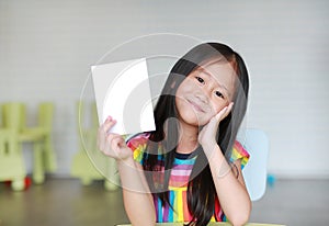 Cute little smiling Asian child girl holding blank white paper card in her hand. Kid showing empty paper note copy space in