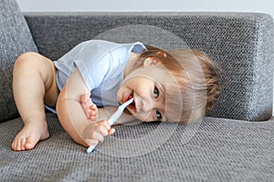 Cute little smiling baby boy holding toothbrush in his mouth and cleaning his first teeth lying in funny pose on the sofa looking