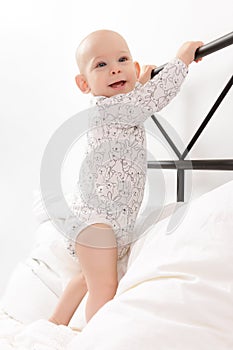 Cute little smiling baby boy infant standing on parent`s bed, holding onto the headboard. First attempts to stand up.
