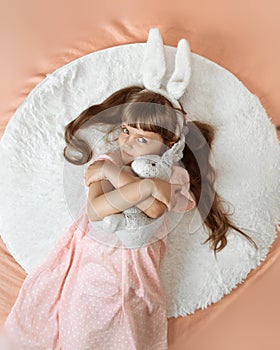 A cute little smiling girl with brown hair is lying on the bed. She has white bunny ears. Baby girl with toy flaffy