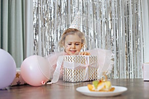 Cute little smiling girl with short curly hair and stars on face in pink poofy dress, cap unwrap gift box near balloons.