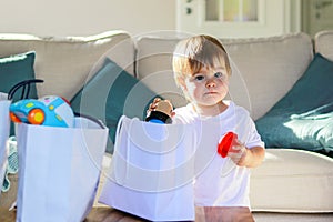 Cute little thoughtful baby boy with funny face expression taking out toys from shopping bags