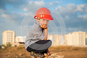 Cute little tired sad kid in orange helmet sitting on background of new buildings and sunset cloudy sky