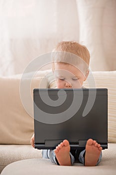 Cute little toddler boy sitting on the sofa with his laptop and looking at the window. Learning with computer