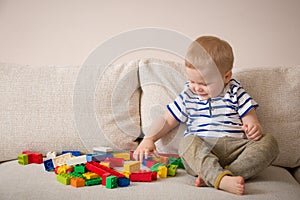 Cute little toddler boy in a striped shirt playing colorful plastic blocks on the sofa indoors. child having fun
