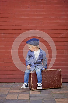 Cute little toddler child, sitting on a vintage suitcase in front of red bricks wall, dressed smart casual
