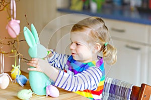 Cute little toddler girl decorating tree and bunny with colored pastel plastic eggs. Happy baby child having fun with