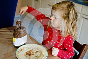 Cute little toddler girl eating homemade fresh crepes or pancakes with chocolate cream for breakfast.