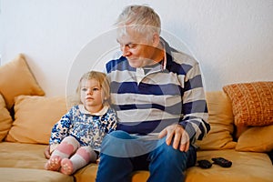 Cute little toddler girl and grandfather watching together tv show. Baby granddaughter and happy retired senior man