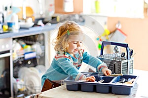 Cute little toddler girl helping in the kitchen with dish washing machine. Happy healthy blonde child sorting knives