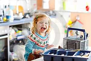 Cute little toddler girl helping in the kitchen with dish washing machine. Happy healthy blonde child sorting knives