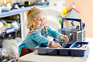 Cute little toddler girl helping in the kitchen with dish washing machine. Happy healthy blonde child sorting knives