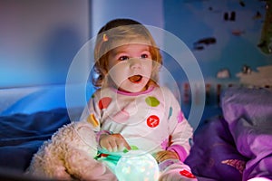 Cute little toddler girl playing with colorful night light lamp before going to bed. Sleepy tired baby daughter in