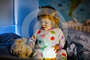 Cute little toddler girl playing with colorful night light lamp before going to bed. Sleepy tired baby daughter in