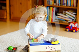 Cute little toddler girl playing at home with eco wooden toys. Happy healthy excited child cutting vegetables and fruits