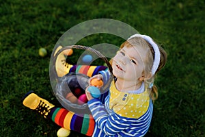 Cute little toddler girl sitting on green grass and wear bunny ears, having fun with traditional Easter colored eggs