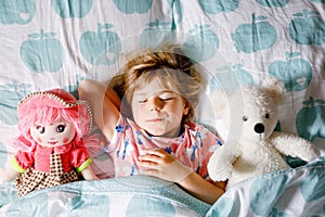 Cute little toddler girl sleeping in bed with favourite soft plush toy doll and teddy bear. Adorable baby child dreaming